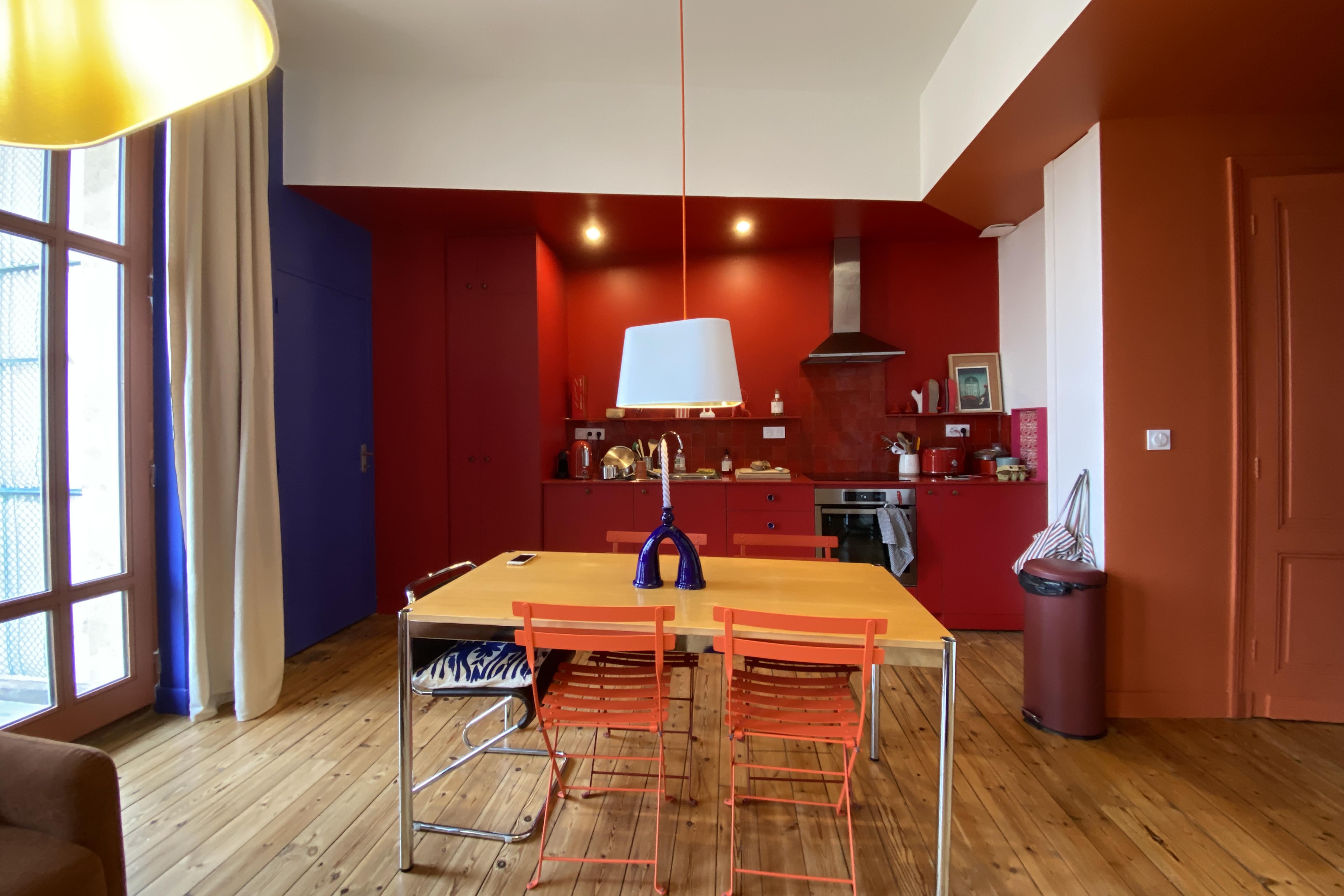 Séjour ouvert sur une cuisine rouge terre cuite et un mur bleu Majorelle, table en bois et chaises orange sous une grande suspension nuage