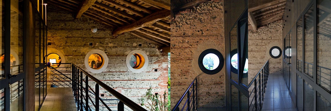Passerelle en mezzanine longeant le mur de galets aux oculi ronds, l'un ouvrant sur la verdure, et boîte de bureaux vitrée à ossature d'acier noir