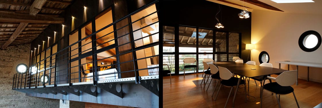 Volume de bureaux en acier et verre noir en surplomb du mur de galets, et salle de réunion au parquet, table sombre et fenêtre ronde sous un puits de lumière