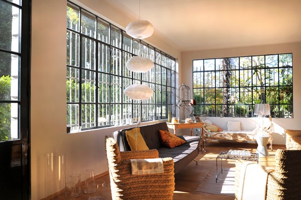 Petit salon sous verrière à structure d'acier noir, suspensions nuage, fauteuils en fibre tressée et canapé face au jardin baigné de lumière
