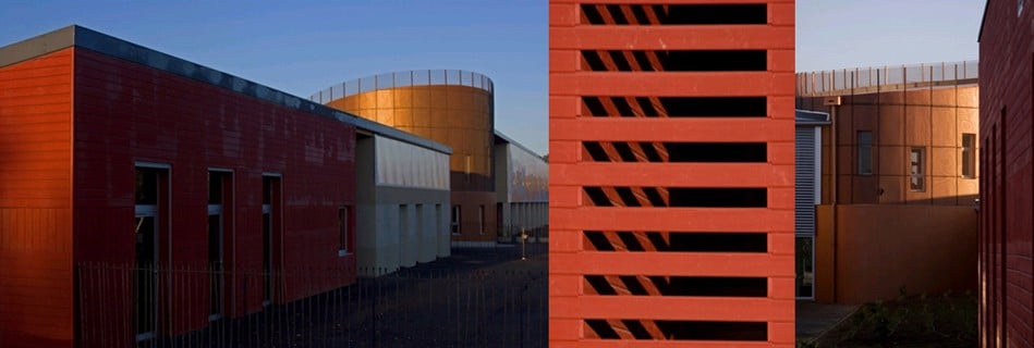 Volumes de l'école en bardage bois rouge au soleil couchant, brise-soleil en lames et rotonde habillée de métal cuivré