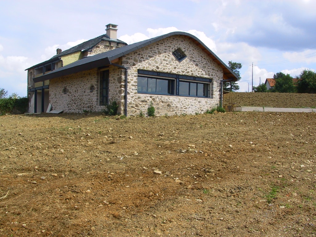 Façade sud de la ferme en pierre, longue baie panoramique en acier noir et toiture cintrée en zinc anthracite, posée à flanc de colline