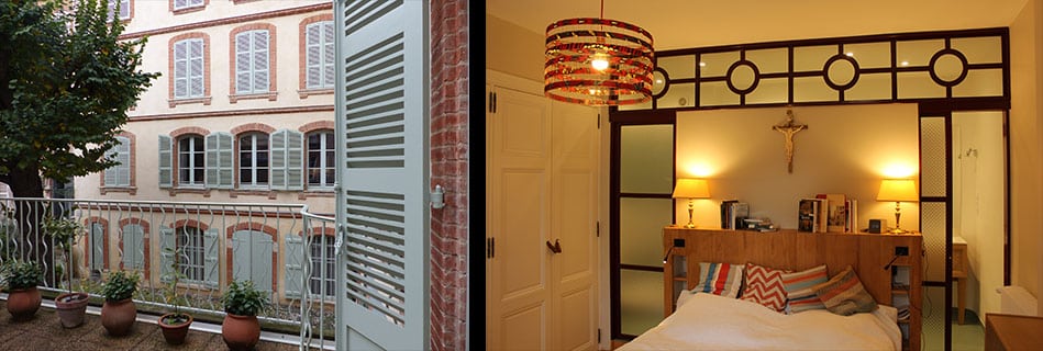 Balcon sur la cour aux façades en brique et volets bois, et chambre séparée par une verrière métallique noire sous une suspension colorée