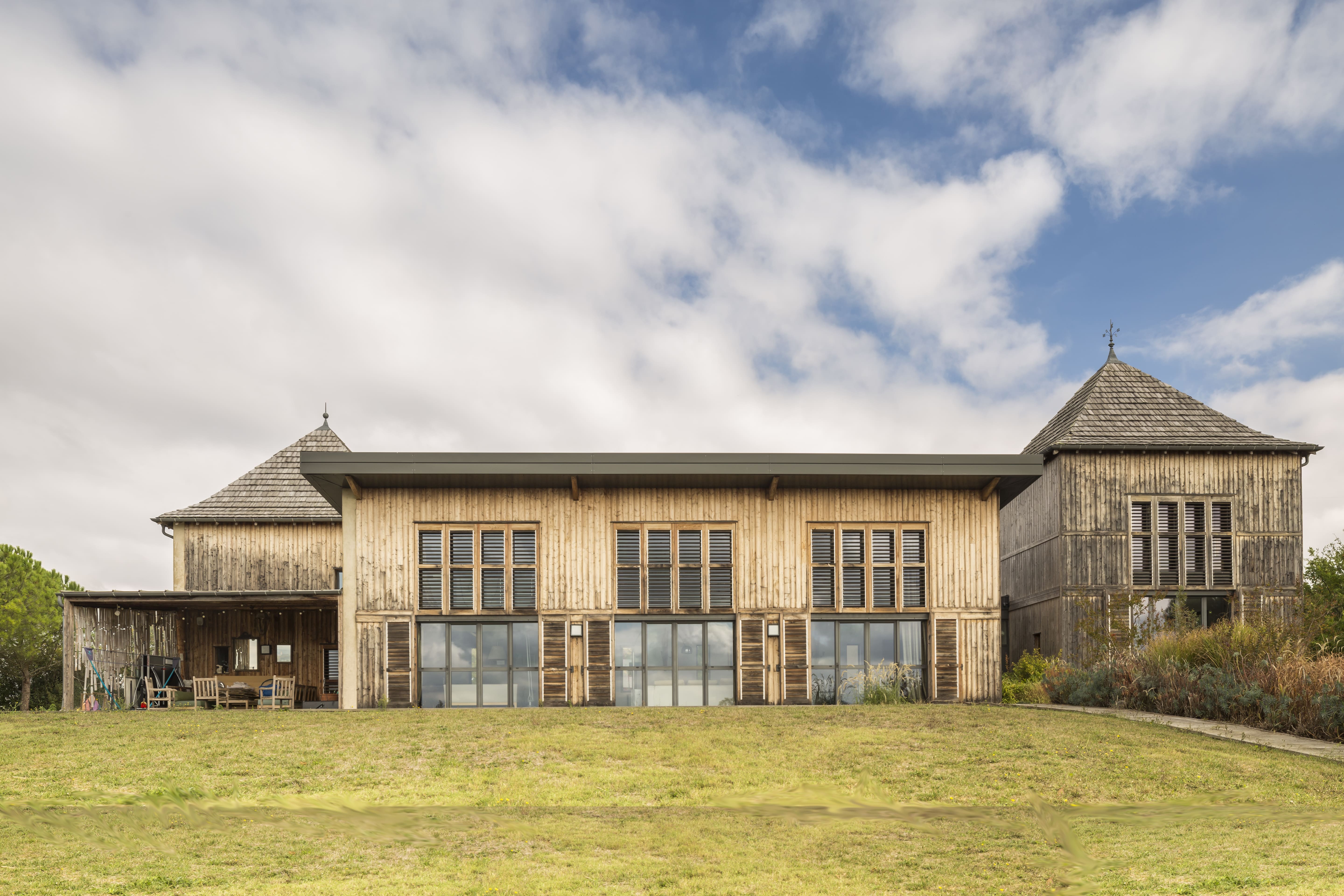 Maison durable au bardage bois vertical, encadrée de deux pigeonniers coiffés de toitures en tavaillons, largement vitrée sur la campagne