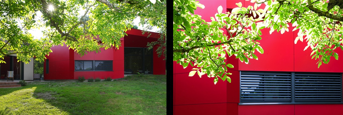 La maison existante à l'enduit béton chaud reliée aux volumes rouges de l'extension, et détail du bardage rouge percé d'un brise-soleil sous les feuillages