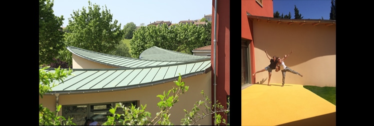 Toiture ondulée en zinc vert au-dessus des murs crème face au village, et patio jaune vif où deux personnes sautent joyeusement le long des façades ocre