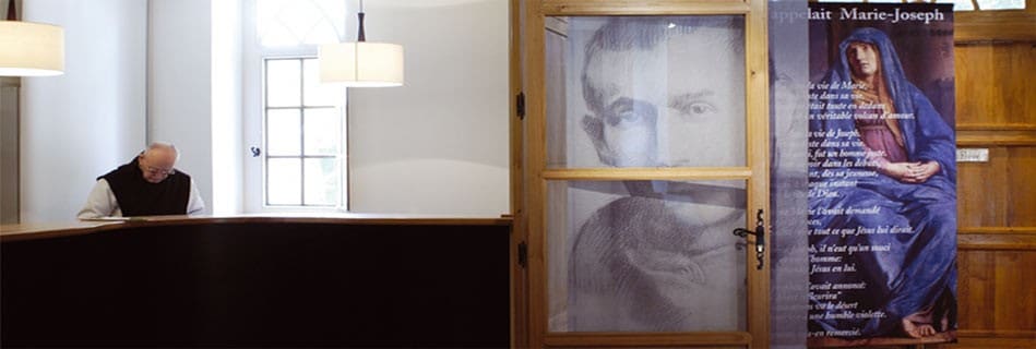 Accueil du monastère avec un moine au comptoir, et porte vitrée au portrait du bienheureux devant un panneau d'exposition