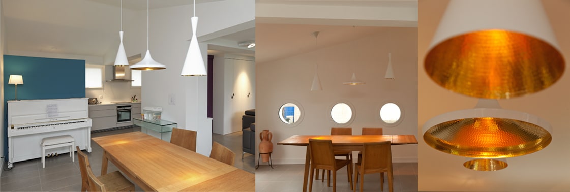 Coin repas sous trois suspensions blanches à intérieur doré, table en bois face à trois hublots ronds, piano droit blanc et cuisine en enfilade
