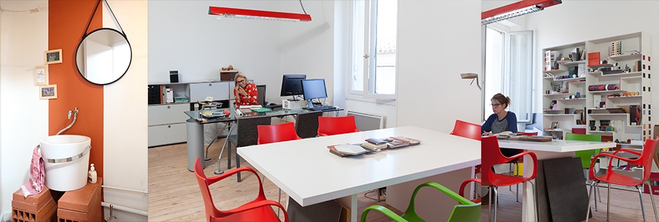 Salle d'eau au mur orange et miroir rond, bureau à la suspension rouge et chaises colorées autour d'une table, et matériauthèque garnie d'échantillons