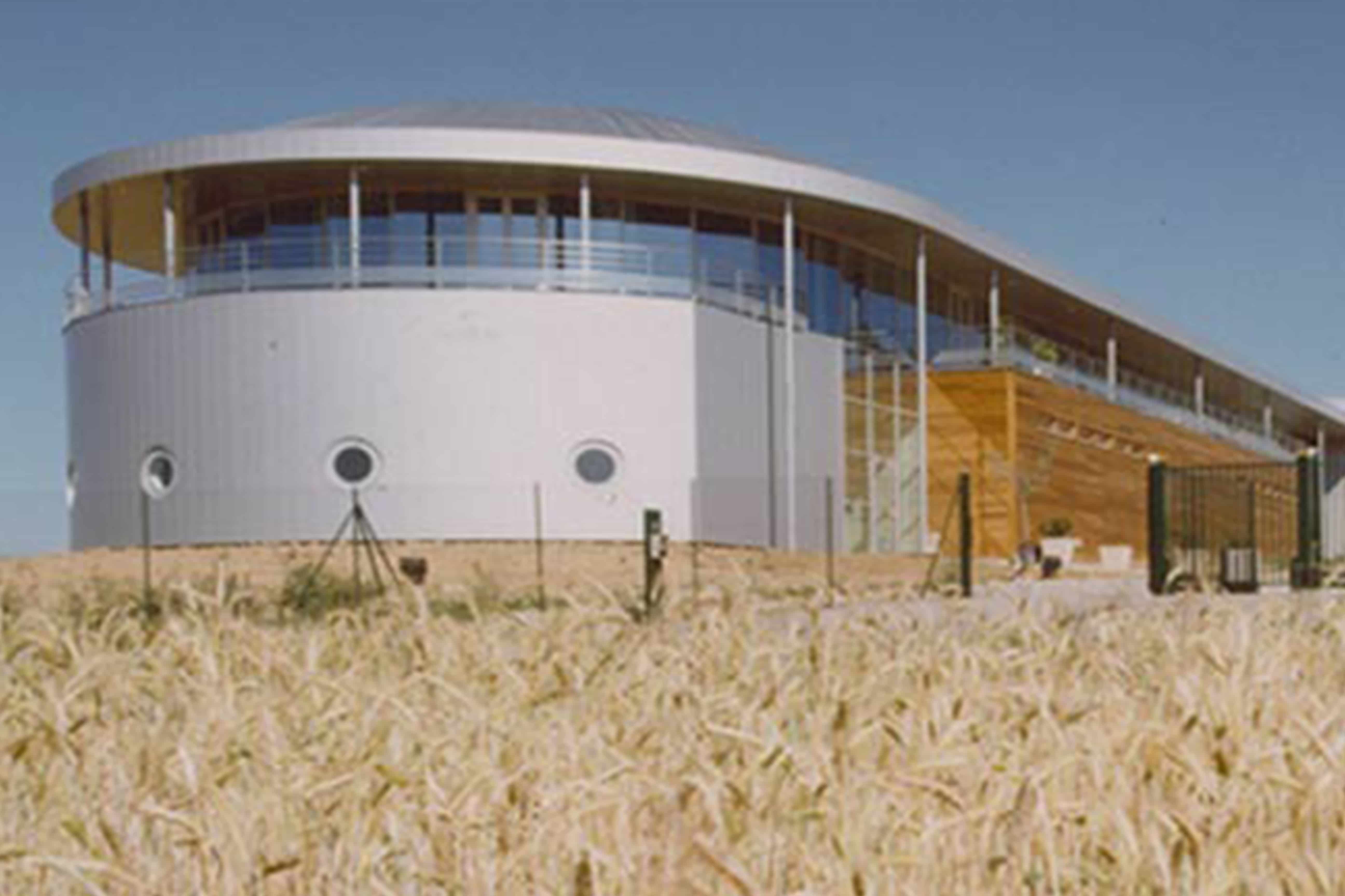 Usine agroalimentaire : bâtiment administratif circulaire blanc à oculi et anneau vitré, relié à une longue halle de production bardée de bois, au milieu des champs de blé